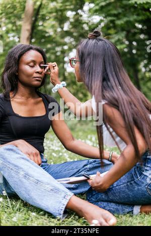 Frau, die Augen schminkt, wenn ein Freund im Park sitzt Stockfoto