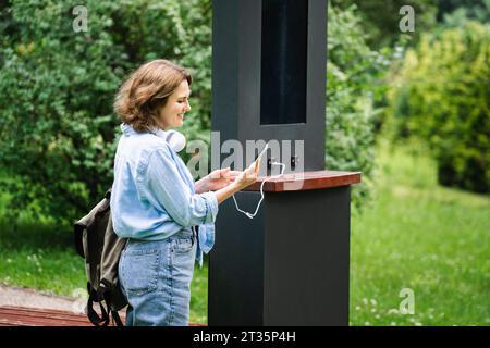 Lächelnde Frau, die ihr Smartphone im Park durch Solarenergie auflädt Stockfoto