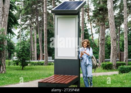 Frau, die ein Handy benutzt und in der Nähe der Solarladestelle im Park steht Stockfoto