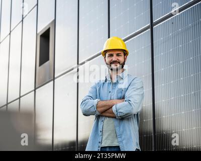 Ein lächelnder Ingenieur trägt einen Helm, der mit gekreuzten Armen vor Solarpaneelen steht Stockfoto