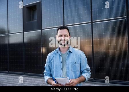 Lächelnder Ingenieur mit Denim-Hemd, der vor Solarpaneelen steht Stockfoto