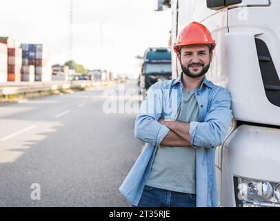 Lächelnder Lkw-Fahrer mit Helm, stehend mit gekreuzten Armen und lehnt sich auf den Lkw Stockfoto