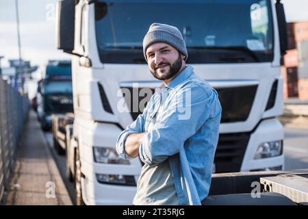 Glücklicher Lkw-Fahrer mit überkreuzten Armen, der am sonnigen Tag vor dem Lkw steht Stockfoto