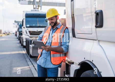 Lächelnder Lkw-Fahrer mit Tablet-PC, der sich auf den Lkw lehnt Stockfoto