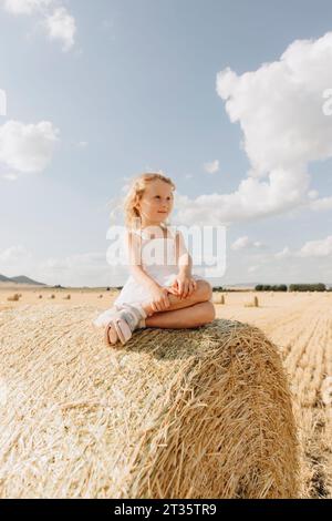 Blondes Mädchen sitzt auf Strohballen auf dem Feld an sonnigem Tag Stockfoto