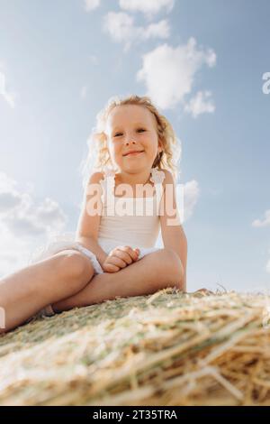 Lächelndes Mädchen, das am sonnigen Tag auf einem Strohballen sitzt Stockfoto