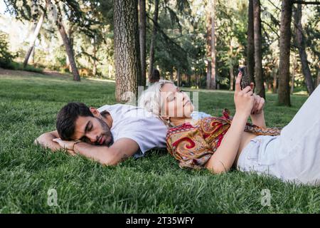 Junge Frau, die mit einem Mann auf dem Gras im Park ihr Smartphone benutzt Stockfoto