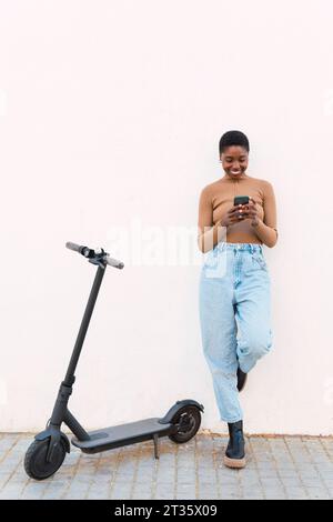 Glückliche junge Frau, die ihr Smartphone benutzt, neben einem elektrischen Roller steht und sich an einer weißen Wand lehnt Stockfoto