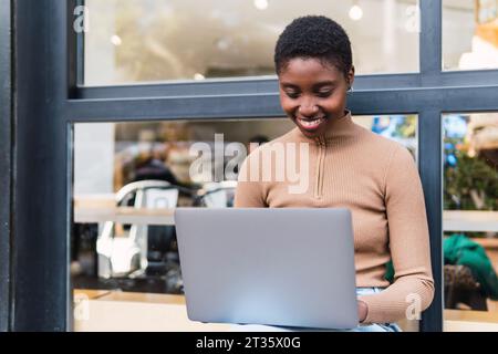 Glückliche junge Frau, die vor dem Café einen Laptop benutzt Stockfoto
