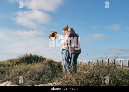 Glücklicher älterer Mann, der mit einer Frau unter bewölktem Himmel einen Hut hält Stockfoto