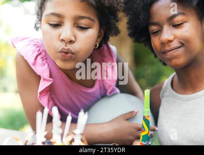 Lächelndes Mädchen mit Schwester, die im Park Geburtstagskerzen bläst Stockfoto