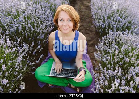 Lächelnde Frau, die mit Laptop im Lavendelfeld sitzt Stockfoto