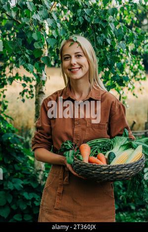 Lächelnde Frau, die einen Korb mit Gemüse im Garten hält Stockfoto