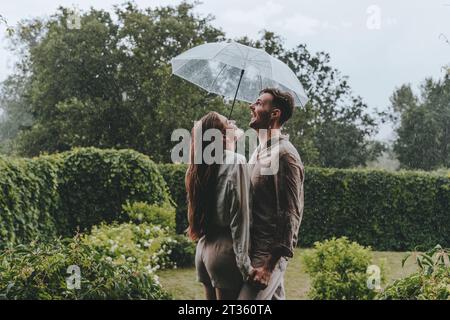 Glückliches Paar, das einen Schirm hält und im Garten den Regen genießt Stockfoto