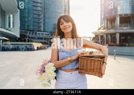 Lächelnde Frau mit Blumenstrauß und Korb am sonnigen Tag Stockfoto