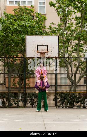 Nicht-binäre Person, die Frau auf Schultern auf dem Basketballfeld in der Nähe des Gebäudes trägt Stockfoto