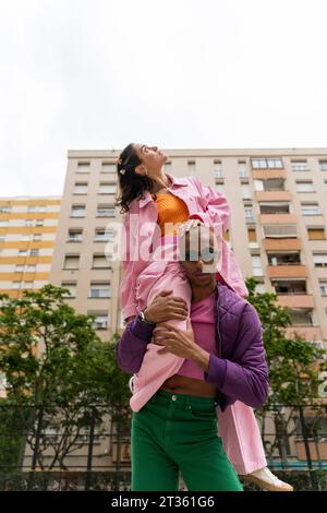 Frau, die auf den Schultern einer nicht-binären Person vor dem Gebäude sitzt Stockfoto