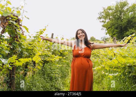 Glückliche schwangere Frau mit ausgestreckten Armen, die neben Pflanzen steht Stockfoto
