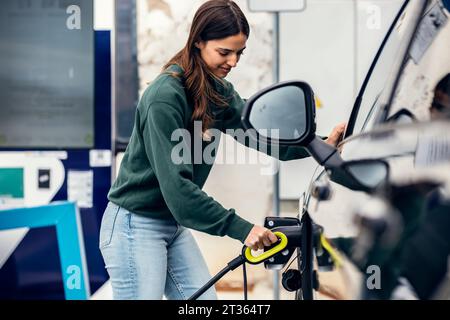 Lächelnde Frau, die das Ladegerät in das Auto an der Ladestation für Elektrofahrzeuge einsteckt Stockfoto