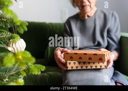 Hände einer älteren Frau, die Geschenke hält und zu Hause neben dem Weihnachtsbaum auf dem Sofa sitzt Stockfoto