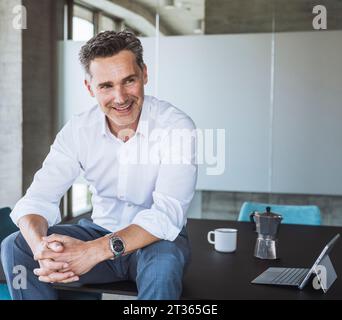 Lächelnder reifer Geschäftsmann, der mit gefesselten Händen auf dem Schreibtisch sitzt Stockfoto
