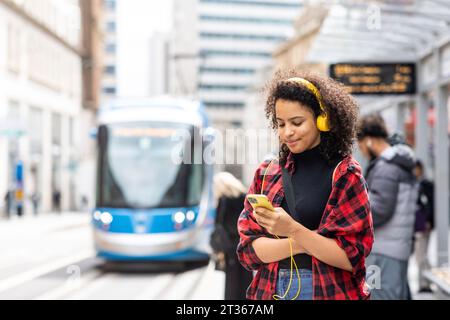 Lächelnde Frau, die Kopfhörer trägt und in der Stadt auf die Straßenbahn wartet Stockfoto