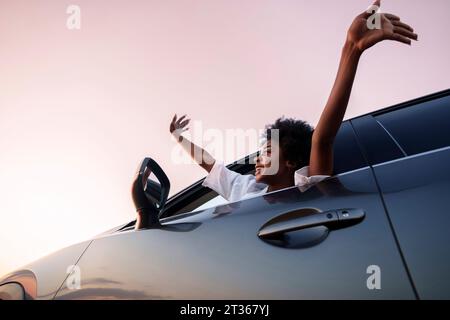 Lächelnde Frau mit erhobenen Armen im Auto unter dem Himmel Stockfoto