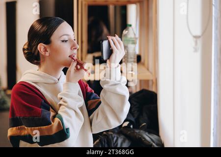 Junge Frau, die Lippenstift aufträgt und im Smartphone schaut Stockfoto
