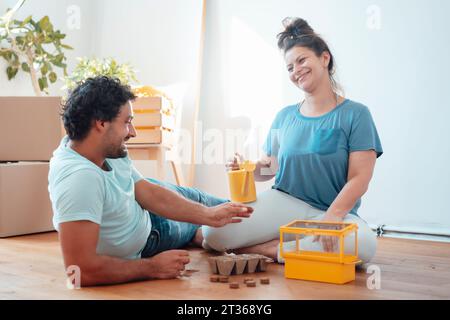 Glückliche Frau, die Samen in Sämling-Tablett pflanzt, während der Mann auf dem Boden liegt Stockfoto