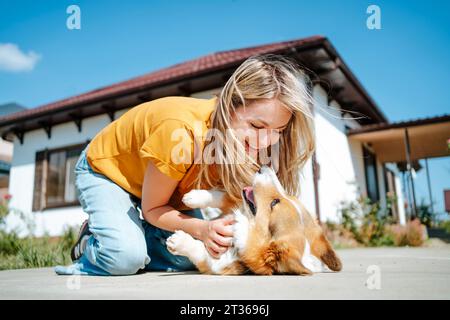 Glückliche Frau, die am sonnigen Tag vor dem Haus mit Corgi-Hund spielt Stockfoto