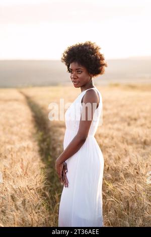 Nachdenkliche junge Frau, die bei Sonnenuntergang im Weizenfeld steht Stockfoto