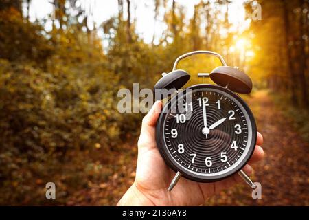 Gersthofen, Bayern, Deutschland. Oktober 2023. Hand hält einen schwarzen Wecker mit 2 Uhr symbolisiert die Winterzeit im Herbstwald *** Hand hält einen schwarzen Wecker mit 2 Uhr als Symbol für Winterzeit in einem herbstlichen Wald Credit: Imago/Alamy Live News Stockfoto