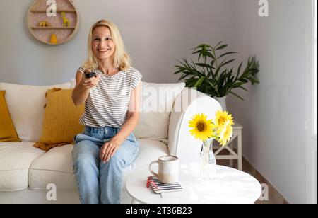 Glückliche blonde Frau mit Fernbedienung im Wohnzimmer zu Hause Stockfoto