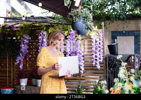 Durchdachter Blumenhändler mit Laptop, der bei Pflanzen im Blumenladen steht Stockfoto