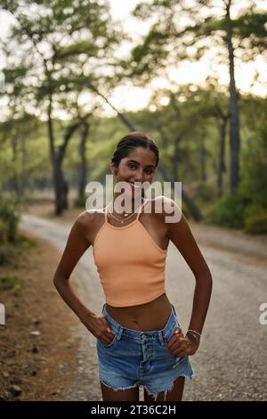 Glückliches Teenager-Mädchen, das bei Sonnenuntergang mit Händen in Taschen steht Stockfoto