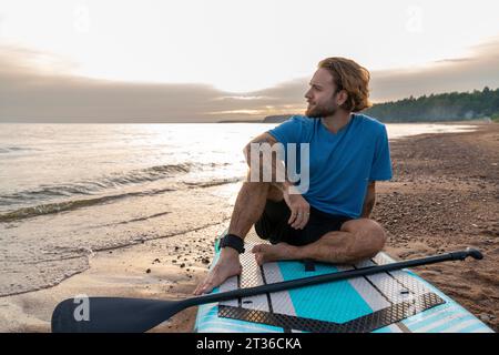 Junger Mann, der am Strand auf dem Paddleboard sitzt Stockfoto