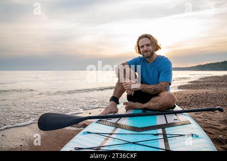 Glücklicher Mann mit Tattoo, der am Strand auf dem Paddleboard sitzt Stockfoto