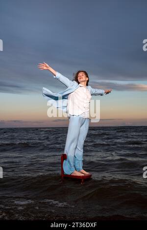 Junge Frau, die auf einem Stuhl im Meer steht, mit ausgestreckten Armen unter bewölktem Himmel Stockfoto