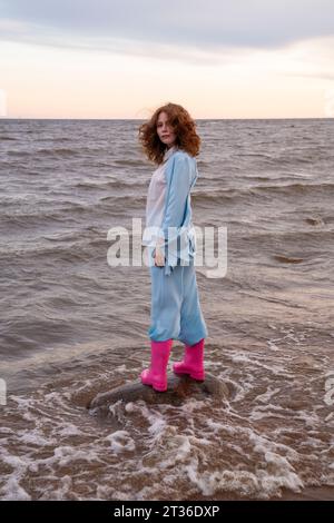 Frau mit rosafarbenen Gummistiefeln, die am Strand auf Felsen steht Stockfoto