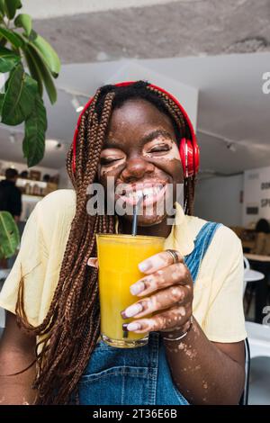 Glückliche junge Frau, die kabellose Kopfhörer trägt und im Café Saft trinkt Stockfoto