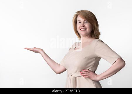 Erwachsene Frau in beigefarbenem Kleid, die in die Kamera schaut. Weibliche Posen, fröhlich lächelnd, mit linkem Arm akimbo Stockfoto