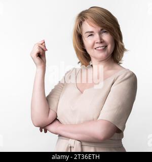 Porträt einer lächelnden glücklichen blonden Frau in beigefarbenem Kleid, die in die Kamera schaut, auf weißem Hintergrund steht. Die Glücksfrau erhob die Hand und schnippt mit den Fingern Stockfoto