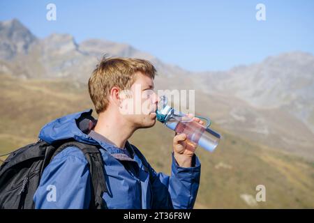 Der blonde Mann trinkt Wasser aus der Flasche in der Nähe des Berges Stockfoto