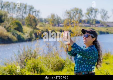 Lächelnde ältere Erwachsene Frau, die Selfie mit ihrem Handy auf dem Land, im Fluss, in Bäumen und in wilder Vegetation in verschwommenem Hintergrund macht, blaue Bluse, bl Stockfoto