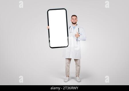 Doktor hält ein riesiges Smartphone und gibt die Daumen hoch Stockfoto