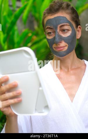 Eine Frau wendet eine graue Tonmaske auf das Gesicht an und schaut in den Spiegel. Stockfoto