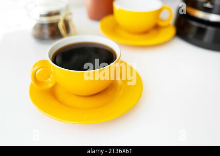 Heißer aromatischer schwarzer Kaffee in einer hellgelben Tasse mit Untertasse steht auf einem weißen Tisch vor dem Hintergrund einer Kaffeemaschine und einer Tasse Kaffeebohnen Stockfoto