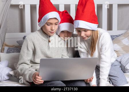 Fröhliche Kinder in roten Weihnachtsmannhüten mit einem Laptop an Heiligabend zu Hause. Jungs und ein Mädchen gratulieren online Verwandten, die auf dem Bett sitzen. Familie x-Mas ev Stockfoto