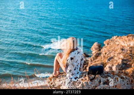 Eine Frau mit langen blonden Haaren, die im Wind fliegen, sitzt auf einem Berggipfel und schaut auf die Meereslandschaft. Stockfoto