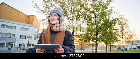 Porträt eines asiatischen Mädchens in warmer Kleidung, sitzt auf einer Bank mit digitalem Tablet und Stift, lächelt fröhlich und zieht bei kaltem Wetter ins Freie Stockfoto